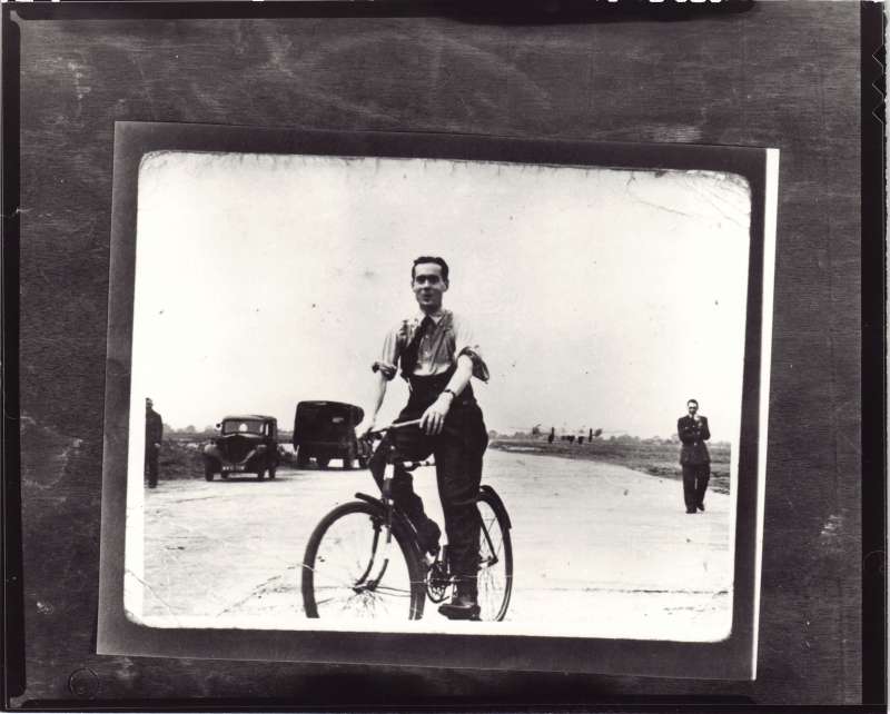 A smiling Leonard Cheshire on a bicycle at an RAF base, with cars and people in the distance