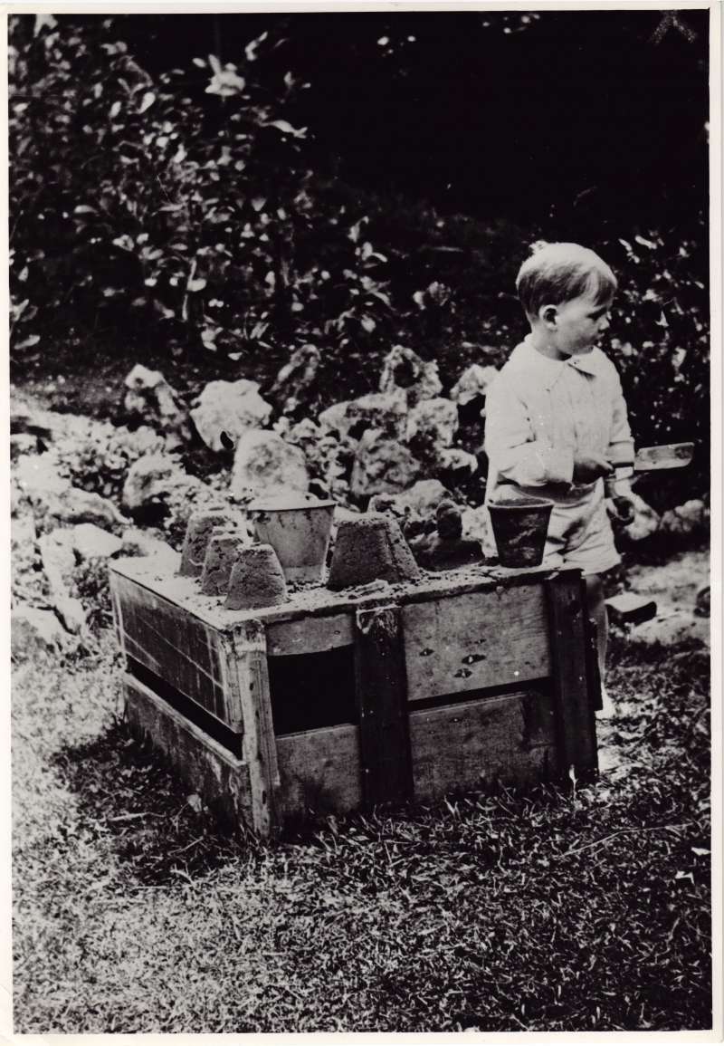A young Leonard Cheshire holding a trowel and playing with sand and buckets in a garden