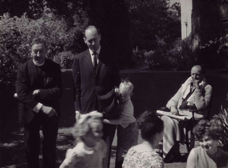 Leonard Cheshire standing outside with others with Jeromy holding on to his leg, and Elizabeth running across the camera
