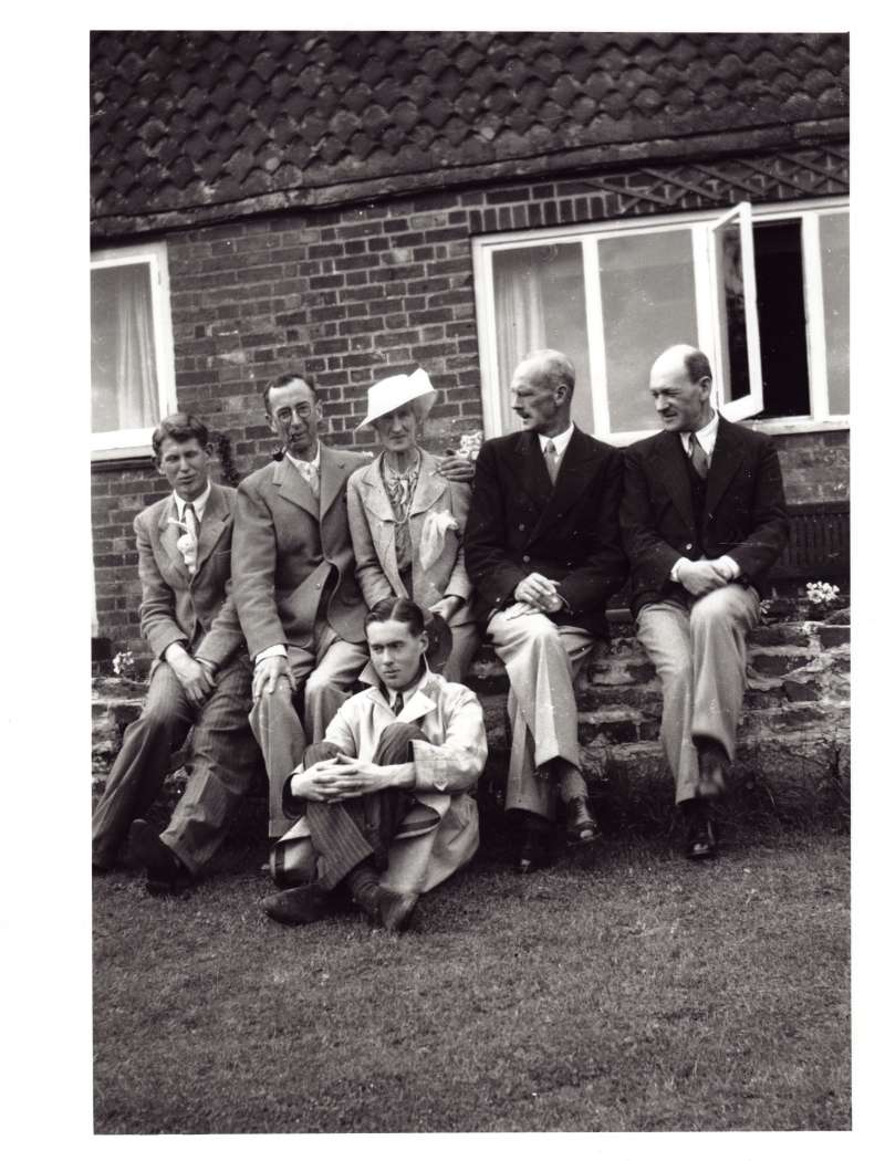 Posed shot of five people sat on a wall outside a house, with a late teenaged Leonard Cheshire sat on the ground in front