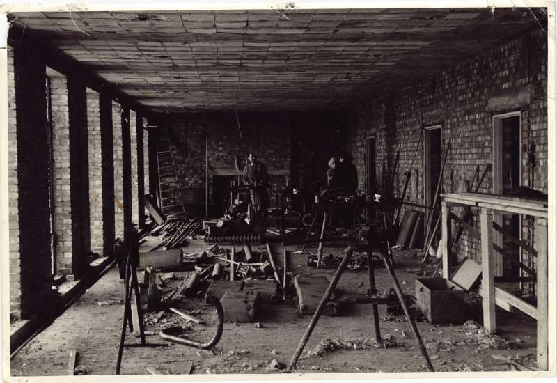 A distant shot of Leonard and his mother inside a half built brick building with building equipment on the floor