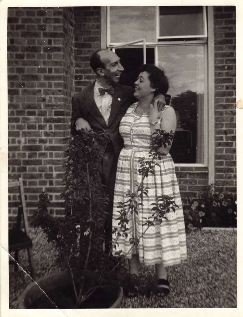 A man standing outside in front of a window with his arm around a lady in a white dress
