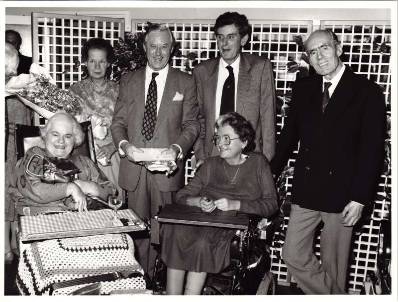 A man and a woman holding gifts standing behind two women in wheelchairs, with Leonard Cheshire standing beside them