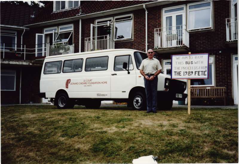 A man standing beside a white minibus in front of a brick building, near a sign saying fete funds will be used to buy the bus