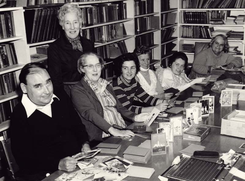 A group of six women and a man sat at a table in a library making christmas cards to sell