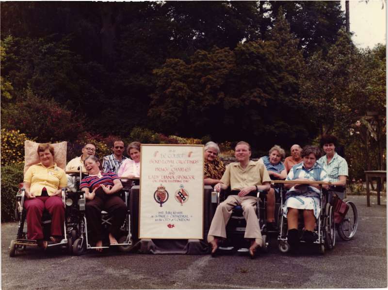 Group photo of several men and women in wheelchairs with a sign celebrating Prince Charles and Diana's wedding