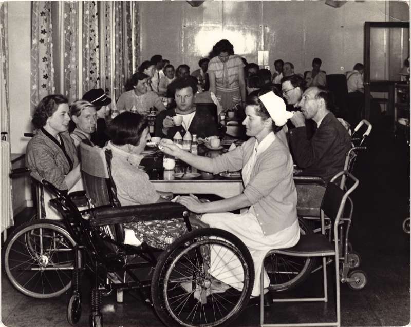 A dining room with several people around tables, and one woman being helped to drink a cup of tea by a nurse