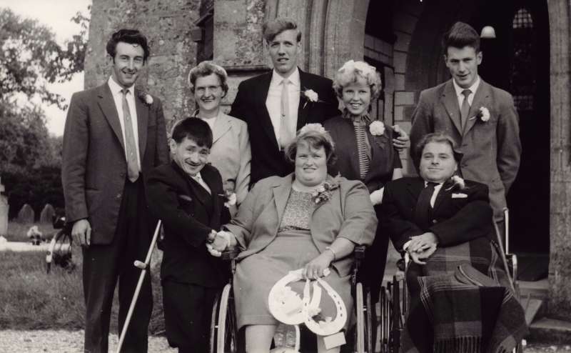 A wedding photo outside a church, the bride and best man in wheelchairs and the groom with crutches