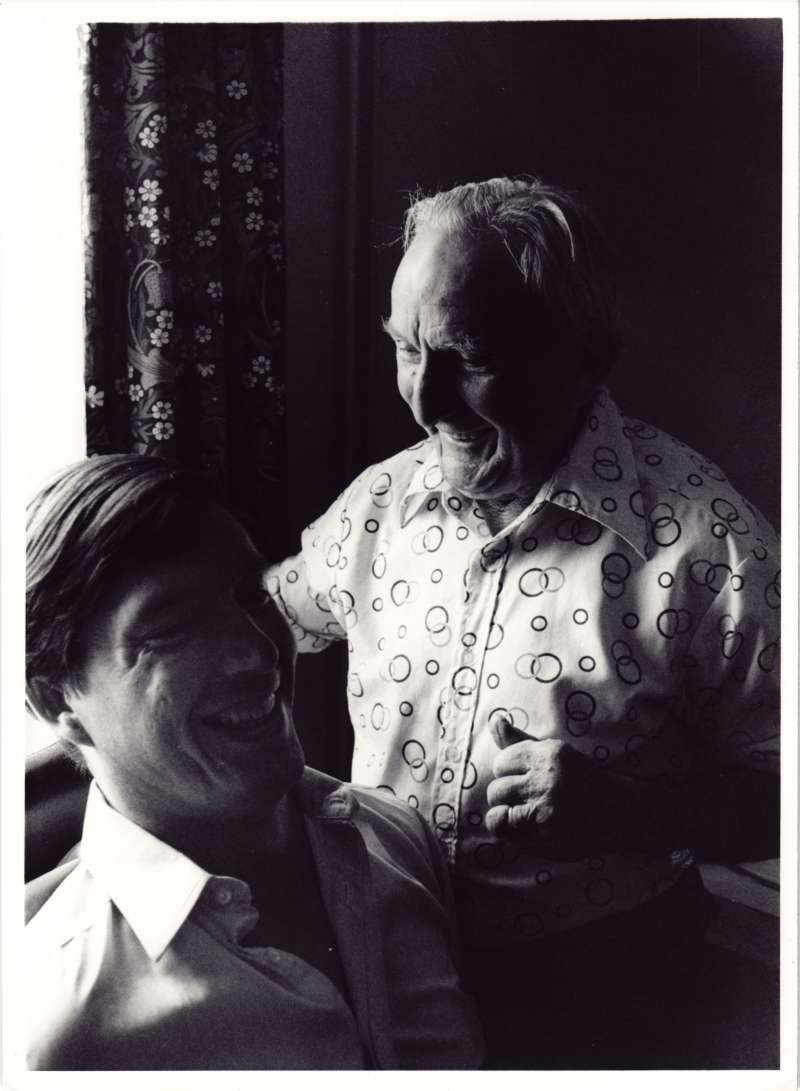 Close up of an older man standing and a younger man sitting, smiling and laughing