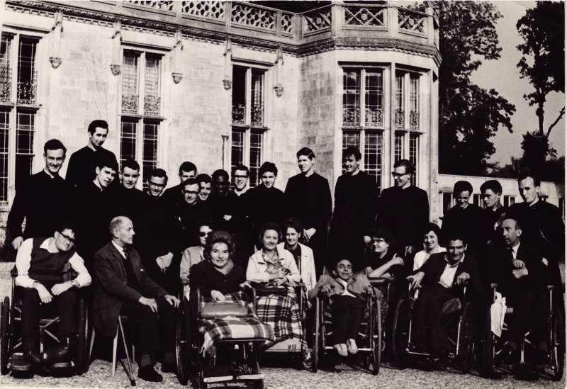 Posed group of several people in wheelchairs and priests in black outfits standing behind, outside at Le Court