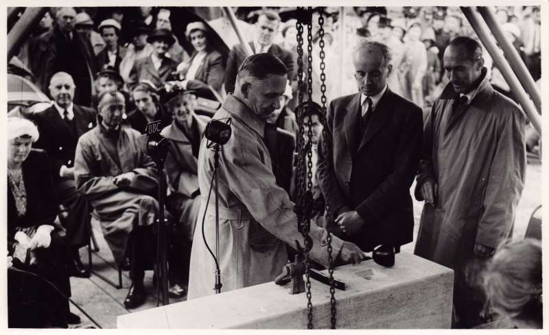A man at a ceremony to lay a large square stone, holding a mallet in his hand, with a crowd behind