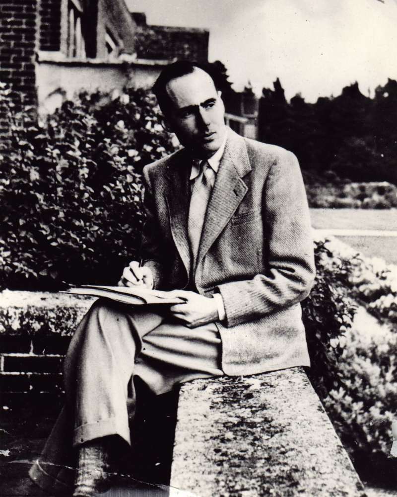 A young man staring into the distance whilst sat on a stone wall with papers on his lap