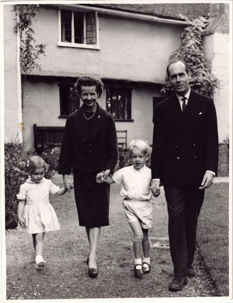 Two young children holding hands with their parents walking in a garden with a house in the background