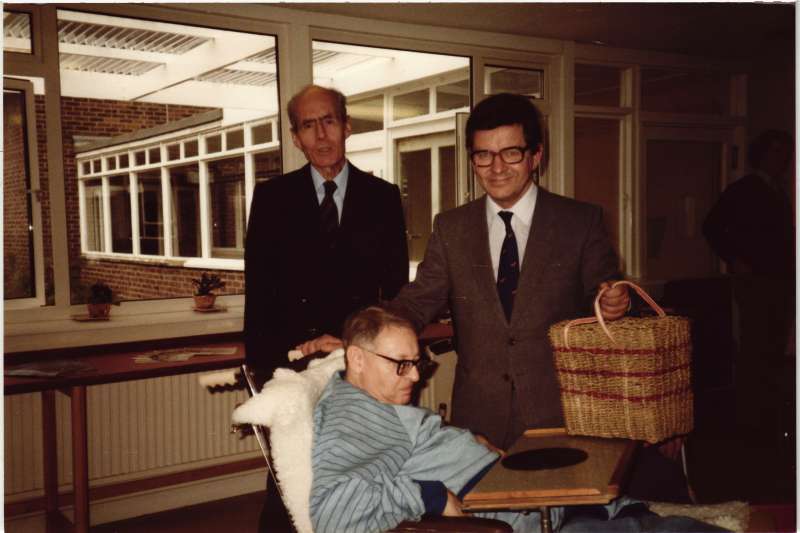 A man in a blue jumper in a wheelchair with Leonard and another man holding a basket standing behind