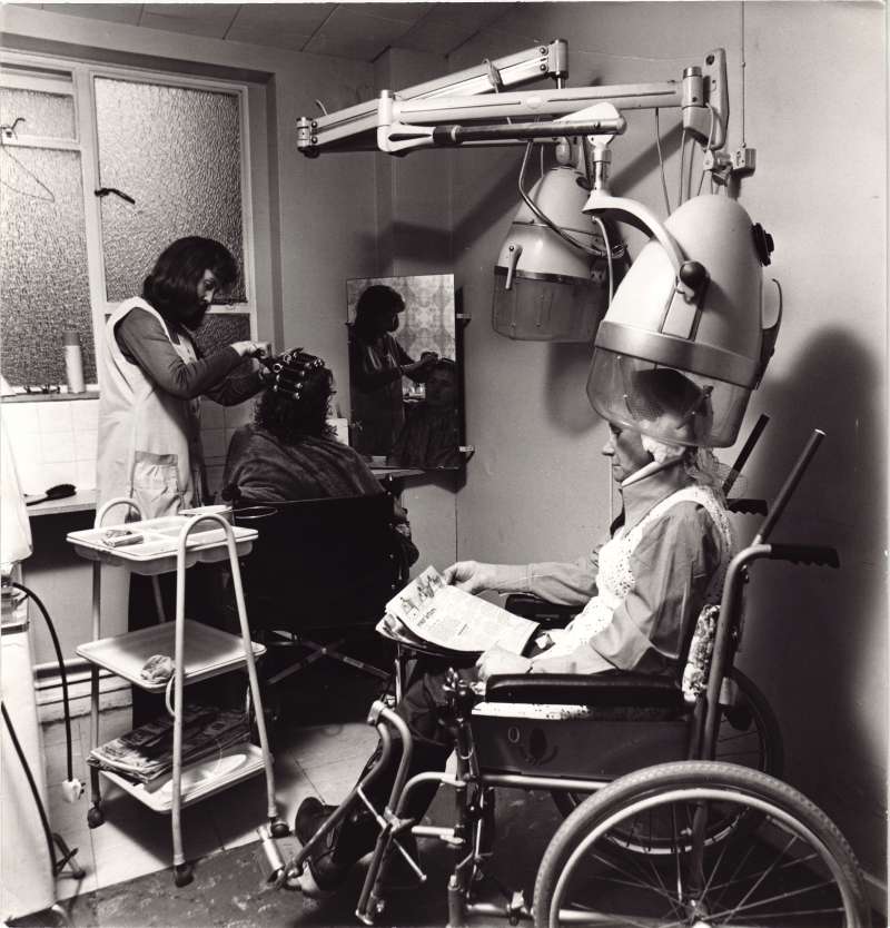 A woman standing putting a lady's hair in rollers, with another lady in a wheelchair sat under a hairdryer