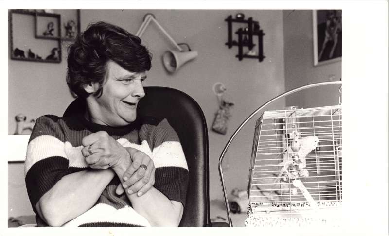 A woman in a bedroom looking excitedly at a budgie in a cage next to her