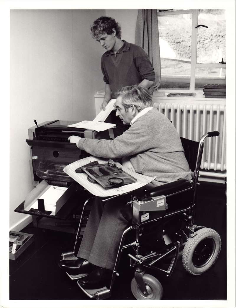 A young man assisting an older man in a wheelchair to use a duplicating machine for newsletters