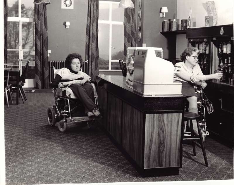 A woman in a wheelchair pouring a drink behind a bar for another lady in a wheelchair