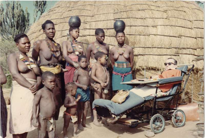 A man in a horizontal wheelchair with several South African women and children outside a woven hut