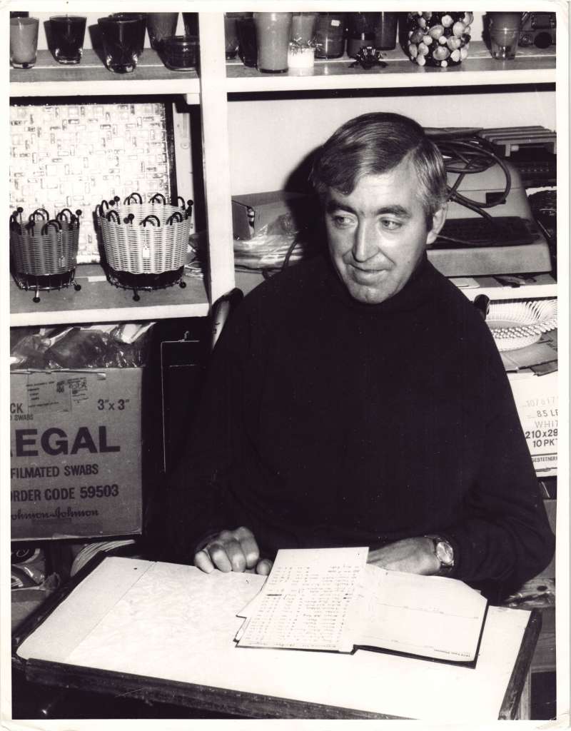 A man in a wheelchair with papers on his lap sat in front of shelves of craft materials