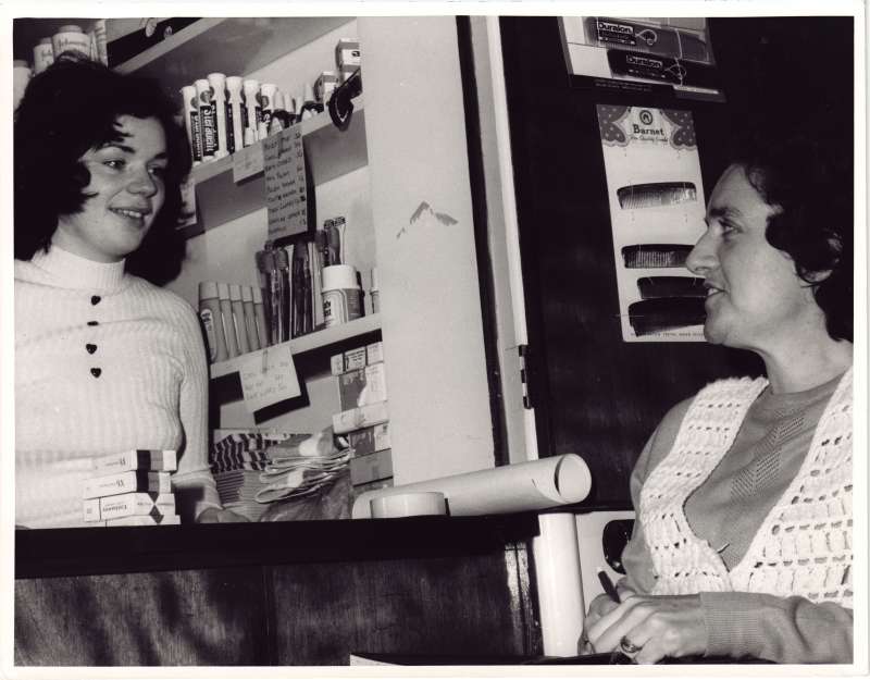A young woman serving another woman in a tuck-shop style shop, with toiletries on the shelves behind