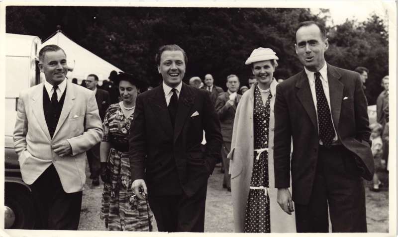 Three men and two ladies walking towards the camera at a fete, including a smiling Leonard Cheshire and Richard Attenborough