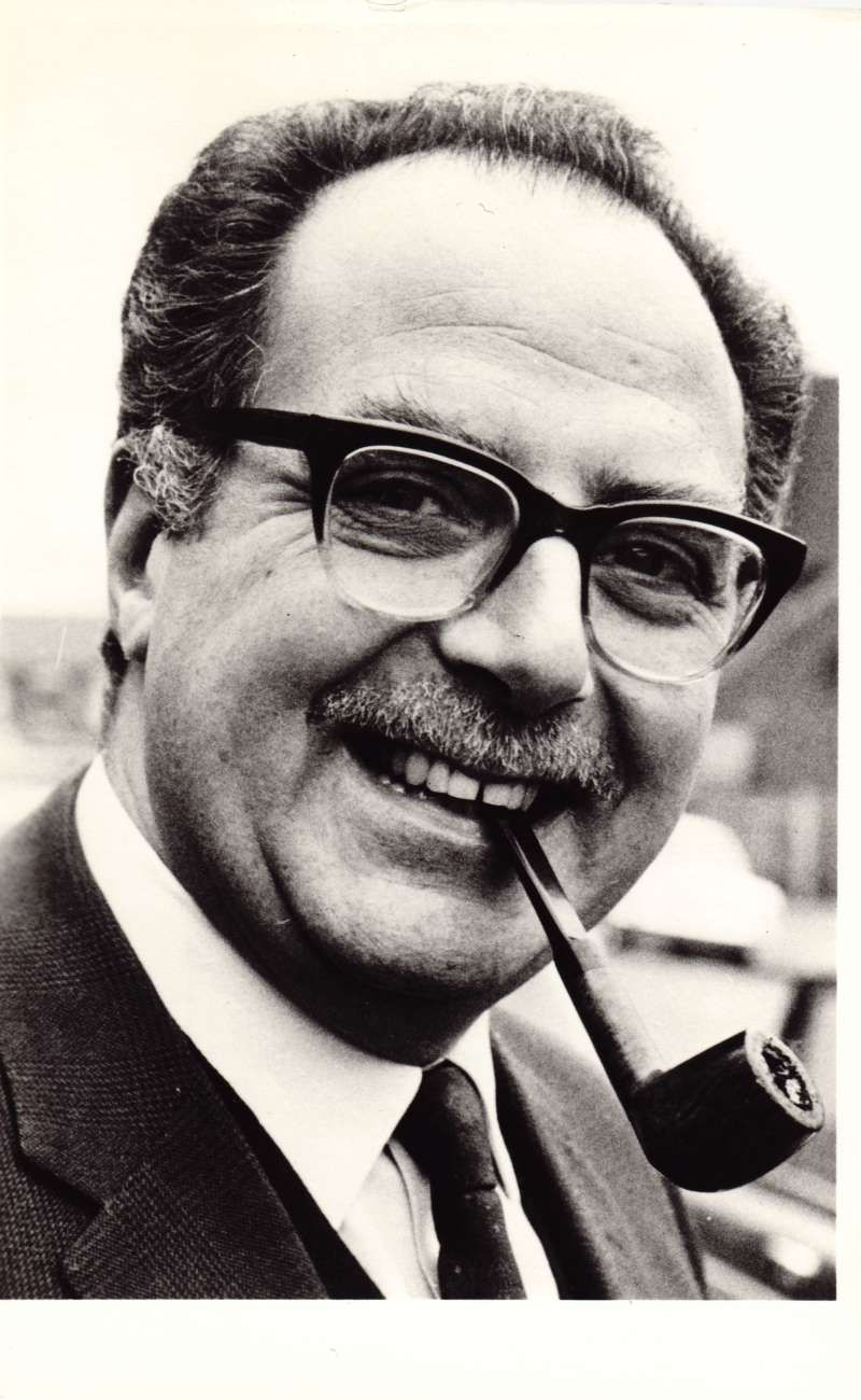A close up of a man in glasses with a moustache and a pipe in his mouth
