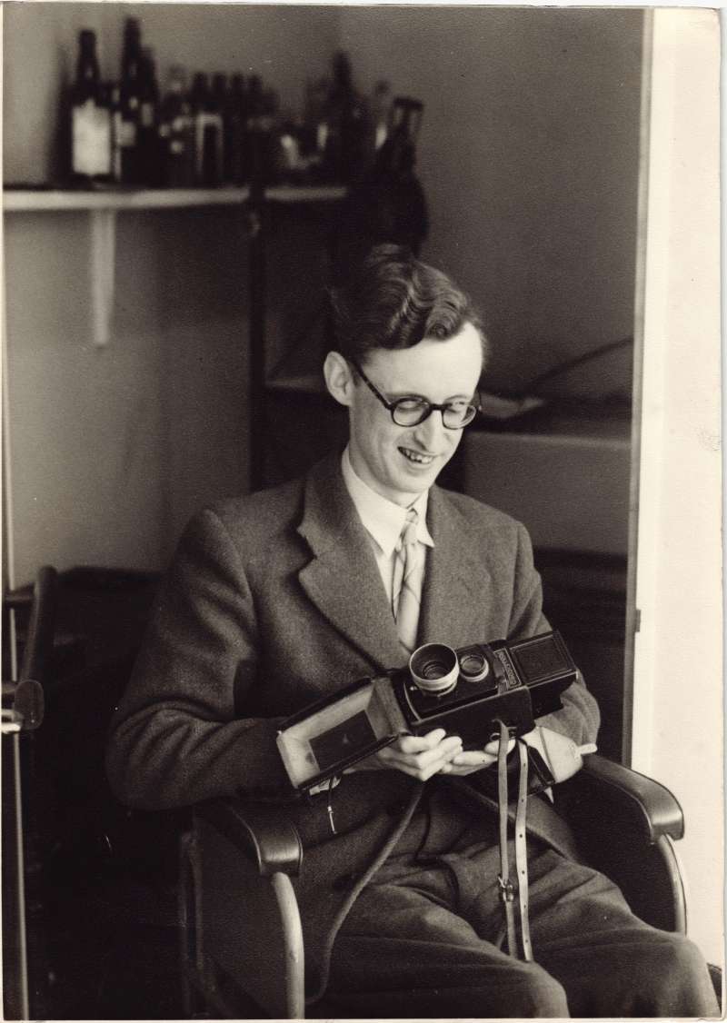 A young man in a wheelchair wearing glasses, holding a old-style camera