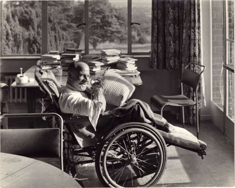 A man in a wheelchair in a light sunny room weaving a basket on his lap with a table covered in books behind