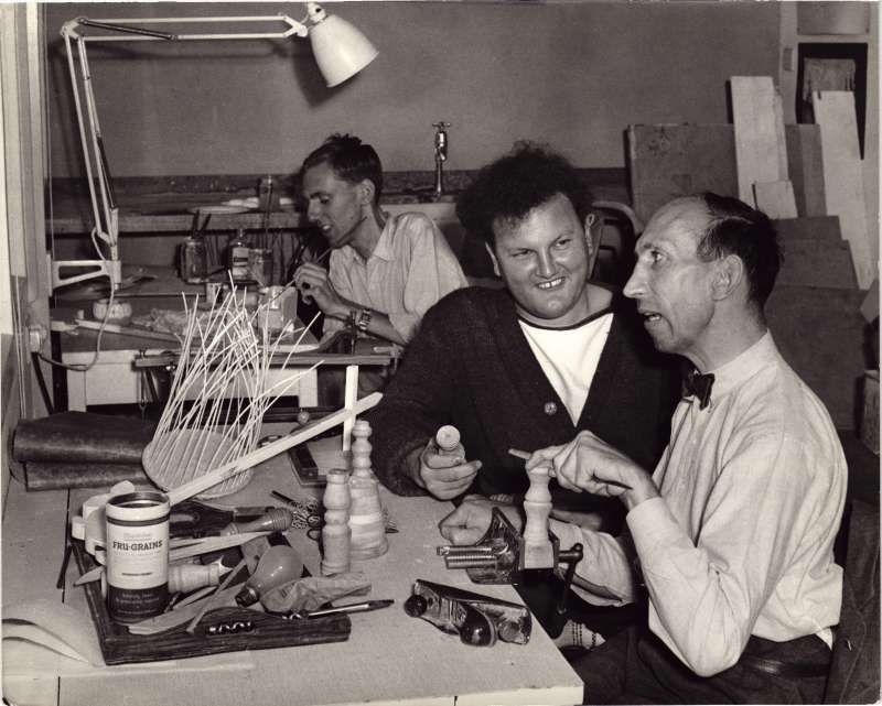 Three men in a workroom doing woodwork and painting
