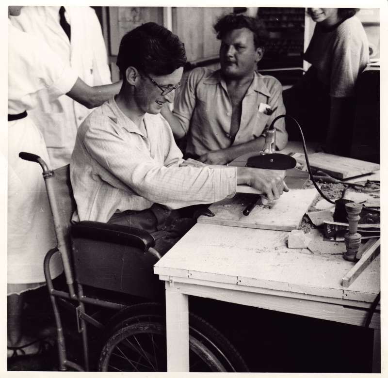 A young man in a wheelchair using a power saw at a workbench with another man and several staff members watching on