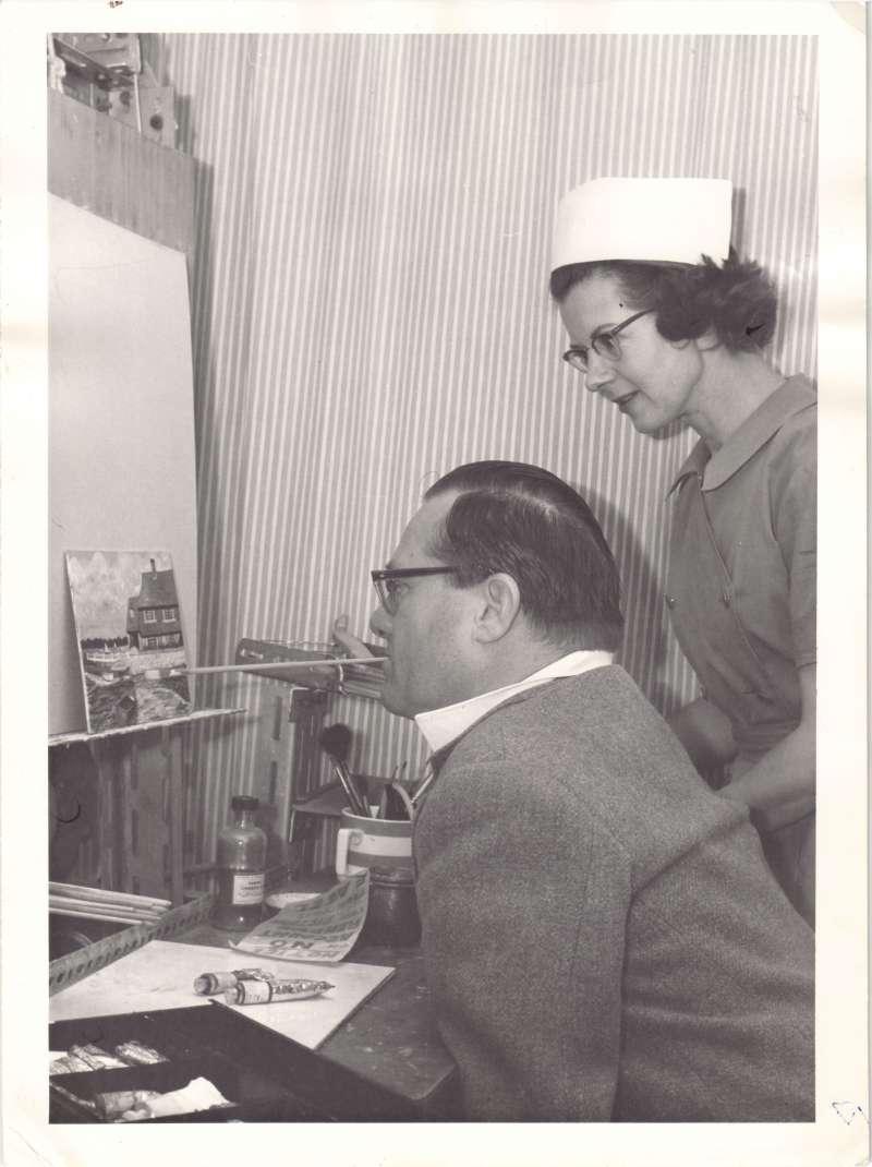 A man painting a picture using a paintbrush in his mouth, assisted by a woman in a nurse uniform