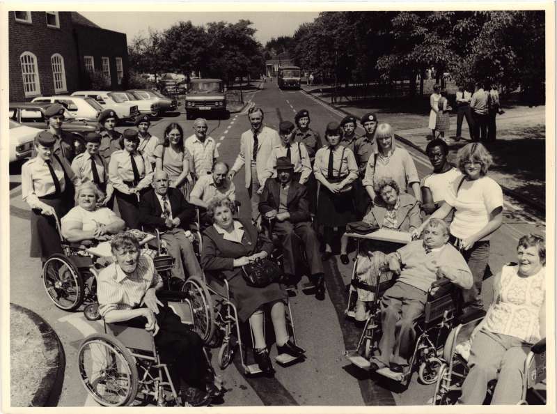 Group shot of several people in wheelchairs and staff members with young people in uniform. Cars and vehicles in the background