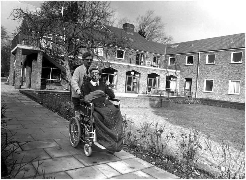 A man in a wheelchair being pushed along by another man along a path in front of the house and gardens