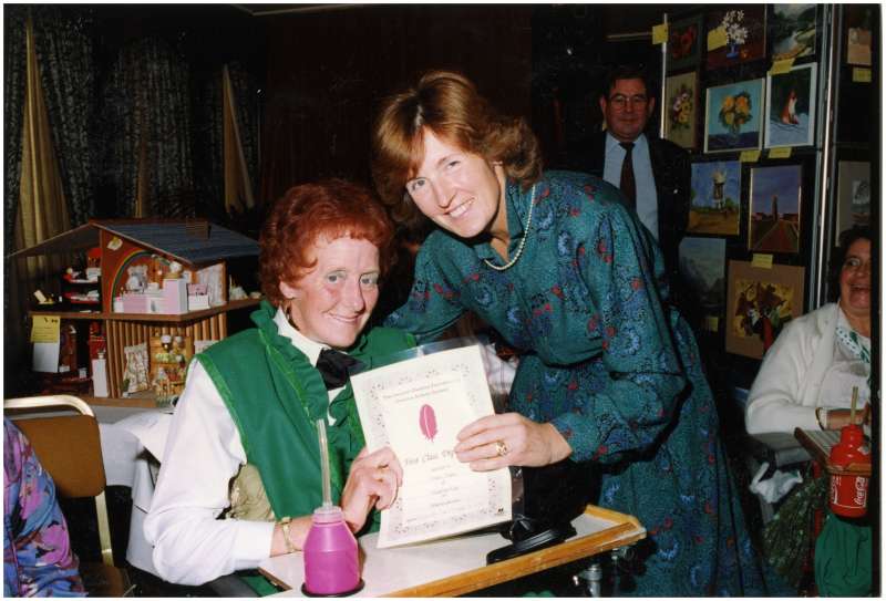 Lady in a wheelchair receiving an award certificate for craft activities from a lady in a green dress