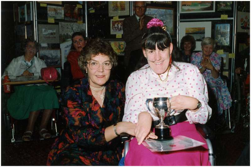 Young lady in a wheelchair wearing a pink and white polka dot blouse receiving a trophy from a lady crouched next to her