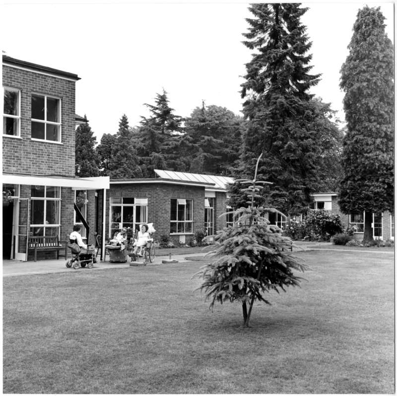 A photo showing the extension at St Cecilia's with lawns and trees. Three people in wheelchairs sat outside