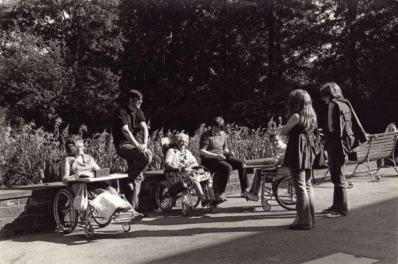 Three residents in wheelchairs talking to four young people outside in the gardens near some benches