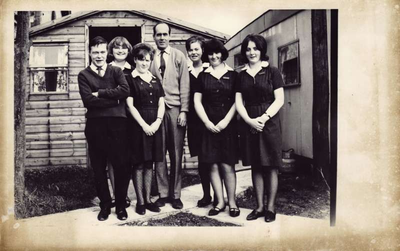 Leonard Cheshire standing in the centre of a posed photo with six young people outside a wooden hut
