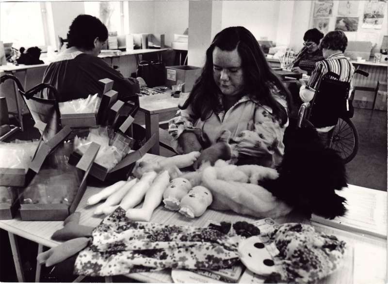Lady at a table making dolls, with three other people in wheelchairs behind at a craft table