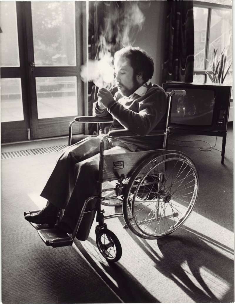 Man in a wheelchair lighting his pipe, caught in a shaft of sunlight through a glass door
