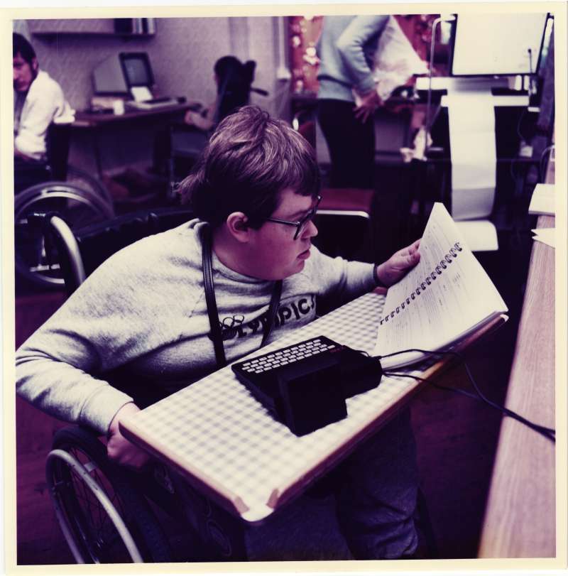 Young man in a wheelchair with a computer keyboard on his lap, holding a book. Others using computers in the background