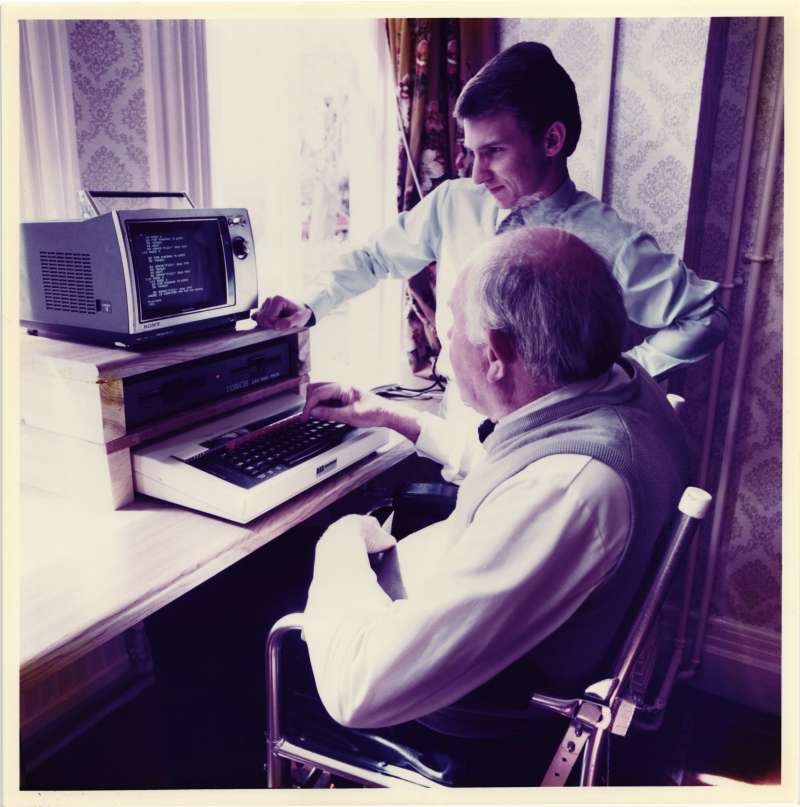 Man in a special chair being helped to use a computer by another man standing beside him