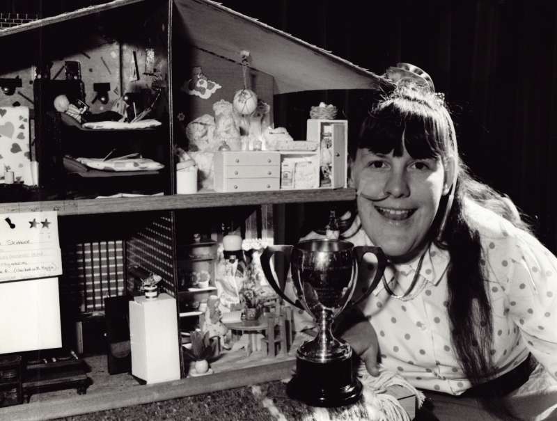Smiling young woman sitting beside a handmade dollhouse, with a trophy next to her