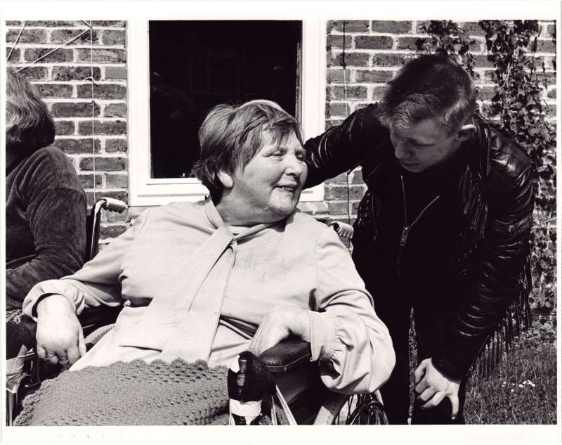Close up of a young man in a leather jacket talking to a female resident in a wheelchair outside a brick building