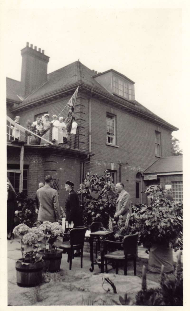 Group of staff standing up in a roof garden overlooking Leonard Cheshire talking with guests below