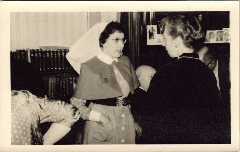 Two ladies, one in a nurse's uniform, talking with others in the background