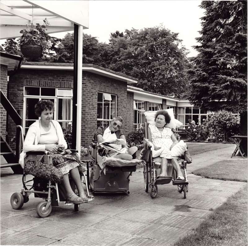 Three people in wheelchairs sat outside. One lady sleeping, one lady knitting, and a man with a newspaper