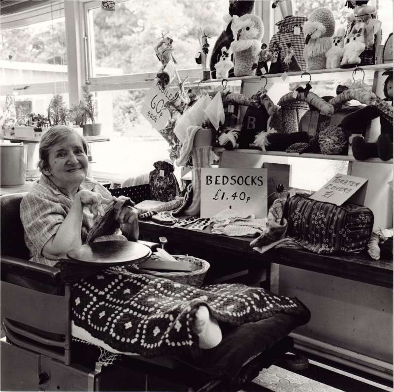 A lady in a wheelchair doing embroidery, with shelves of embroidered dolls, coat hangers and socks next to her