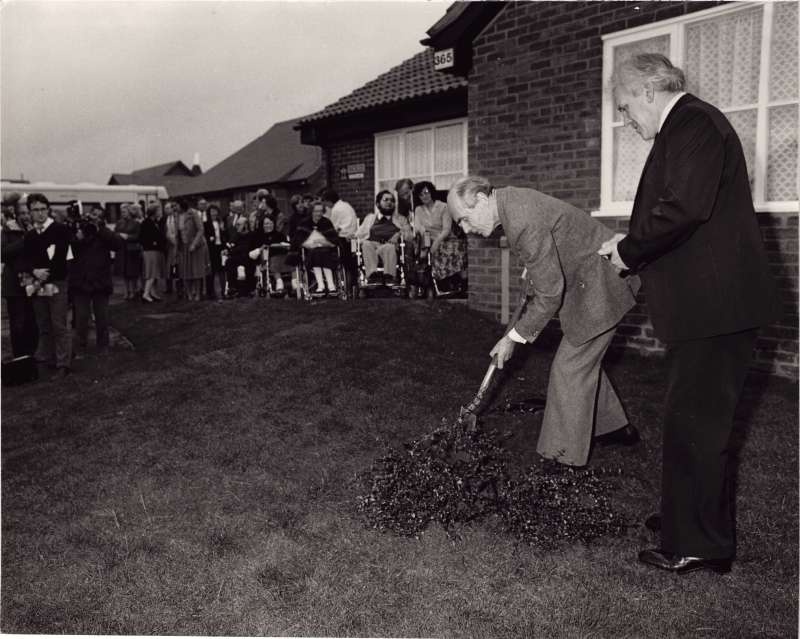 Leonard Cheshire with a shovel turning soil outside new bungalows with staff and residents looking on